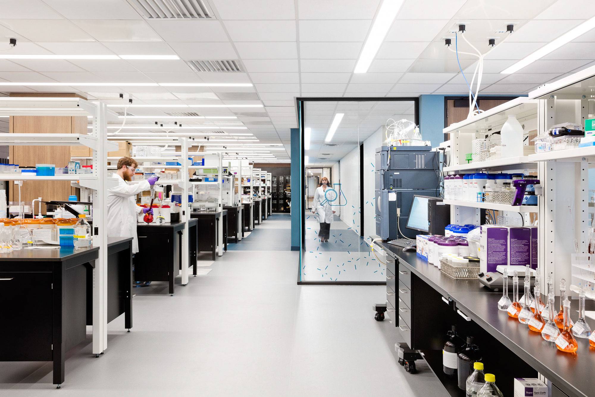 A person in a white coat in a room with shelves and equipment.